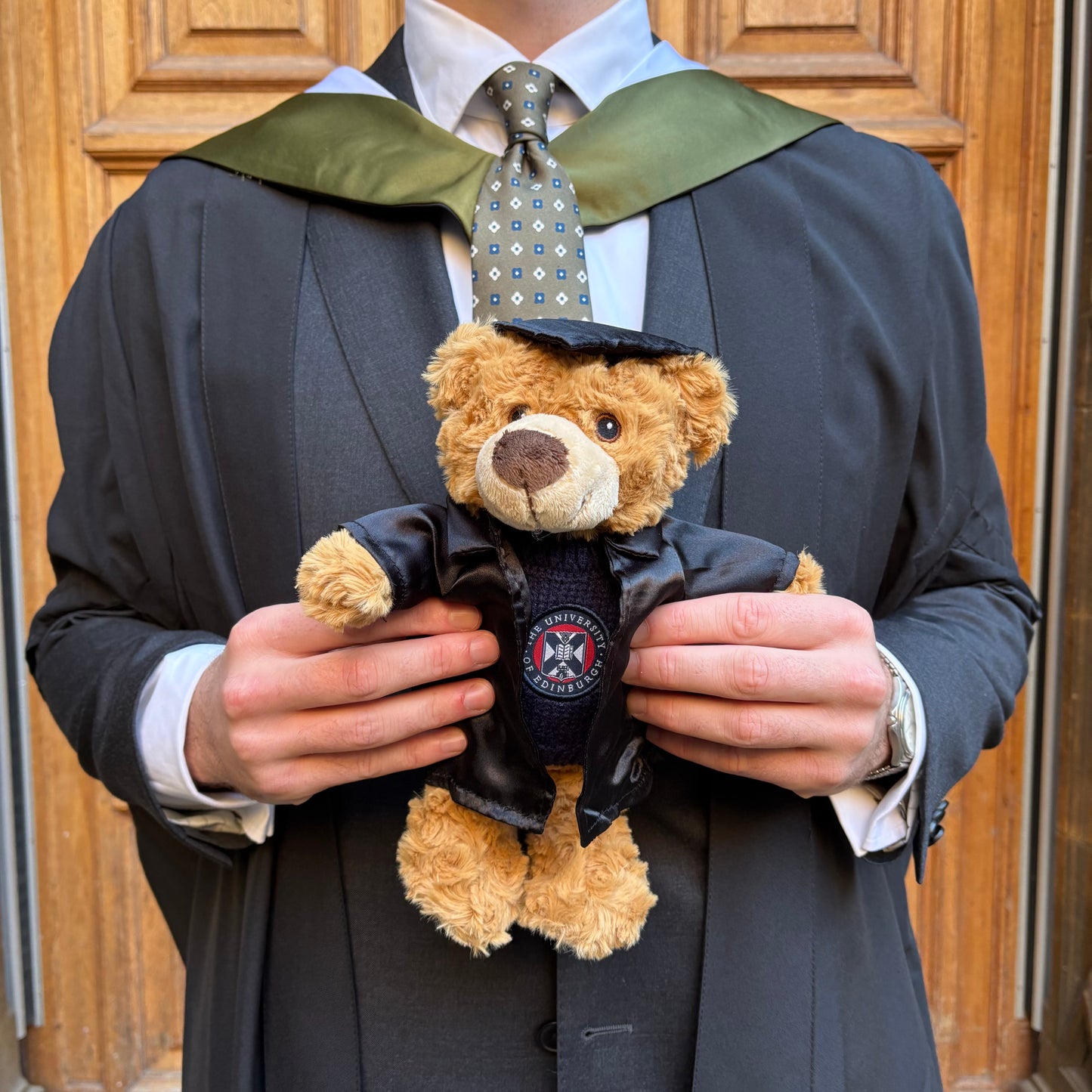 Person in academic gown holding a teddy bear also in academic attire against a wooden door background