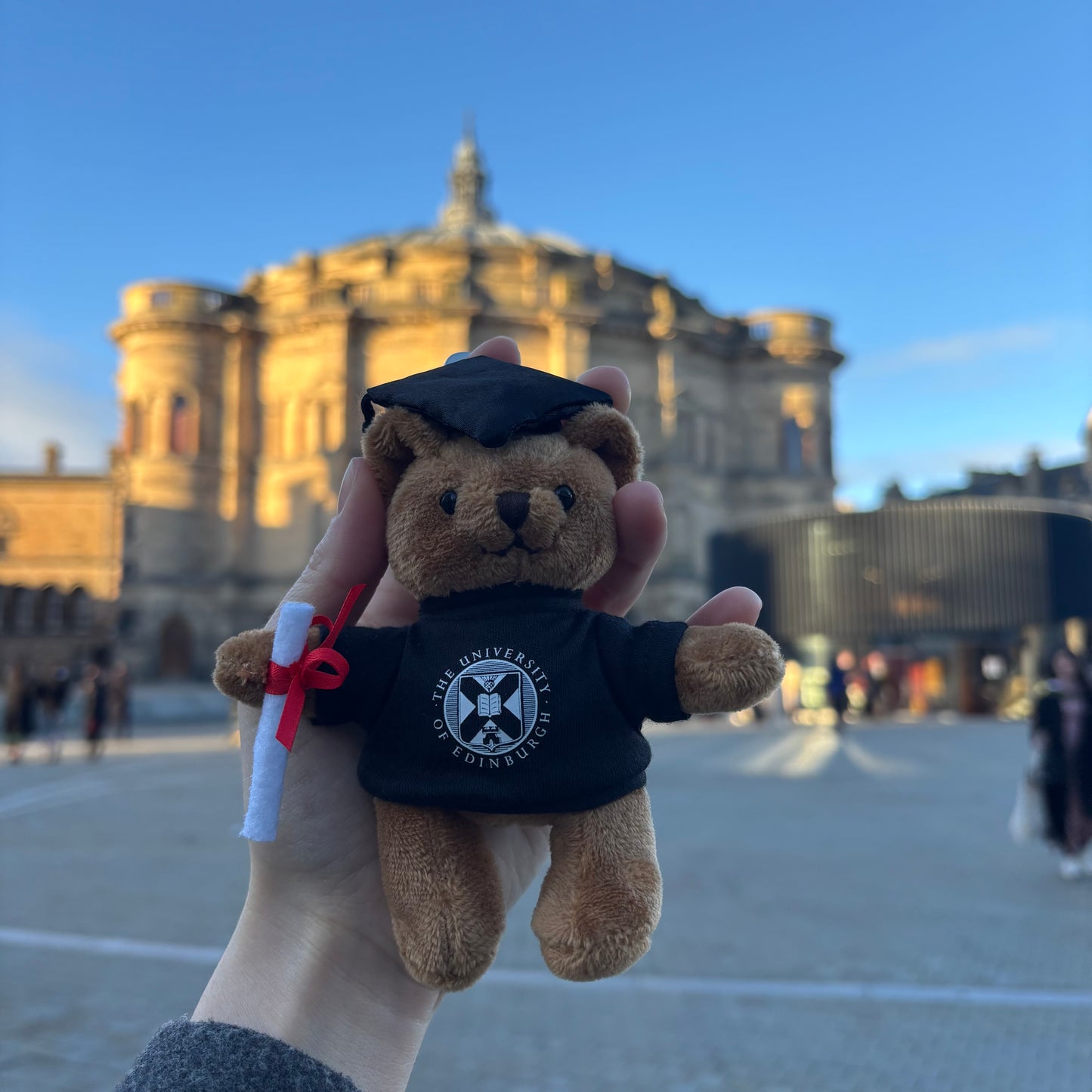 Teddy bear keyring wearing a graduation cap and University of Edinburgh gown with a blurred background of McEwan Hall.