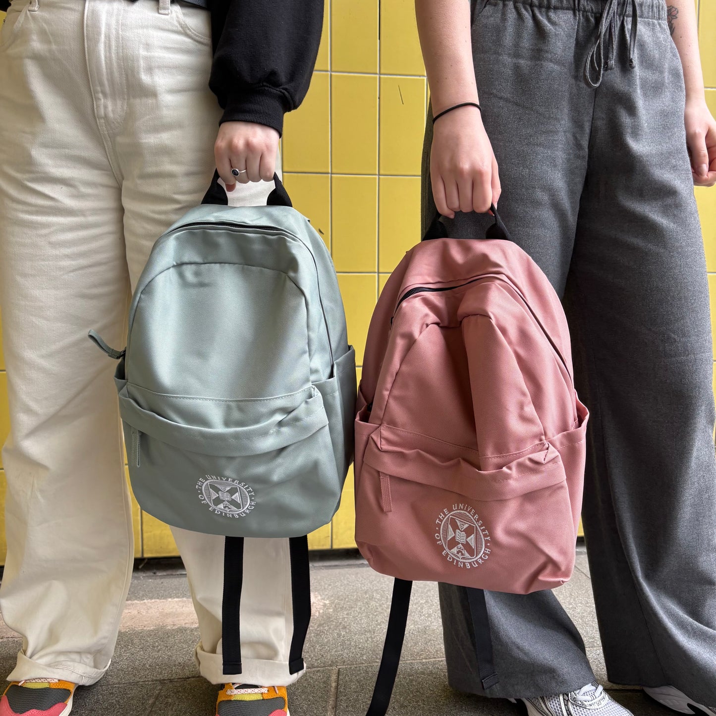 Models carry the Mini Recycled Crest Backpack in dusty green and rose pink. Both bags feature the University crest embroidered in white.