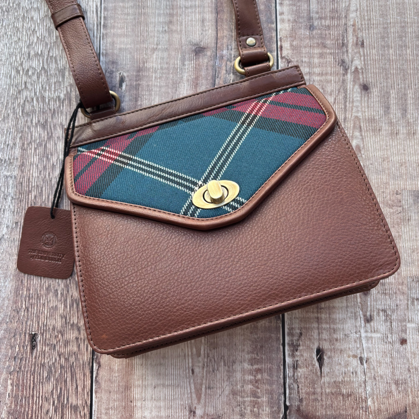 Pictured is the front of the Tartan and Leather Crossbody bag. The bag is brown leather with a tartan flap. The bag features a metal closure made of gold hardware.