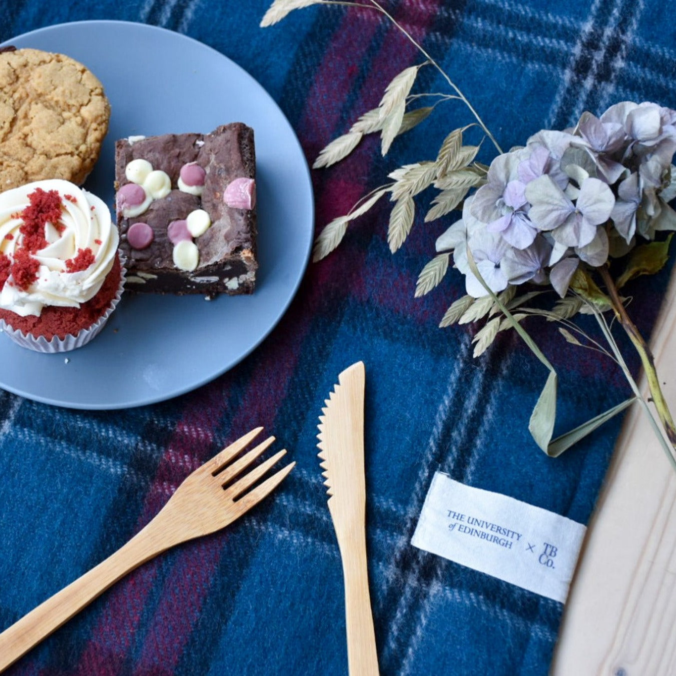 picnic blanket with wooden cutlery, flowers and a plate of cakes