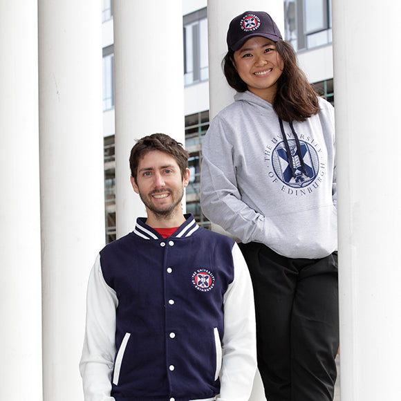 Two models wearing University merchandise. The model on the left is wearing a Varsity Style Baseball Jacket while the right hand model is wearing a Large Crest Two Tone Hoodie in navy and our Embroidered Crest Cap.