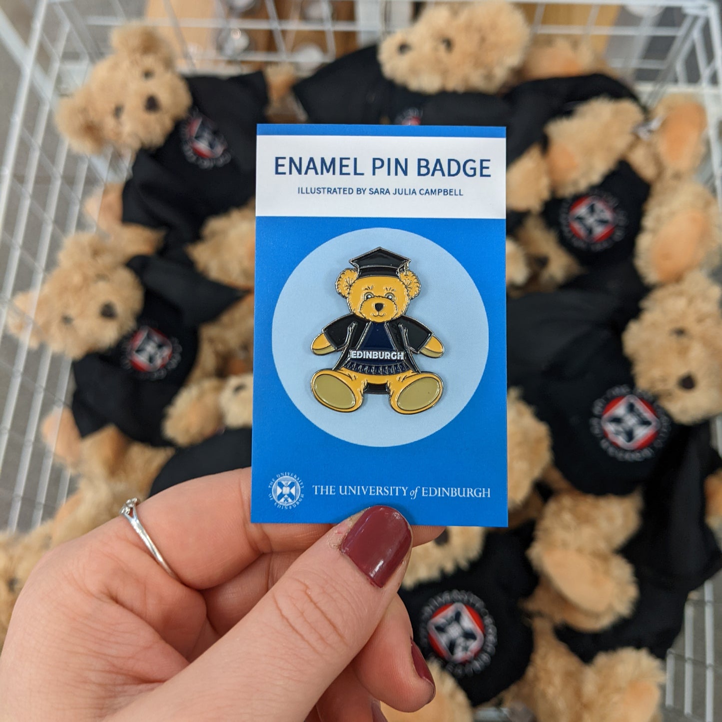 Graduation outfit Edinbear pin badge in front of a basket of plush Edinbears. 