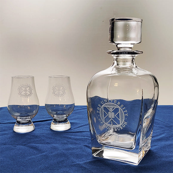 Two University crest branded whiskey tasting glasses next to a Crystal decanter with carved glass stopper featuring the University crest.