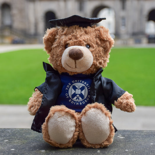Edinbear Recycled Plush sitting on a concrete bench in front of the quad in Old College. He is wearing a black graduation robe, black graduation hat, and a navy blue tee shirt with the University of Edinburgh's crest in white.