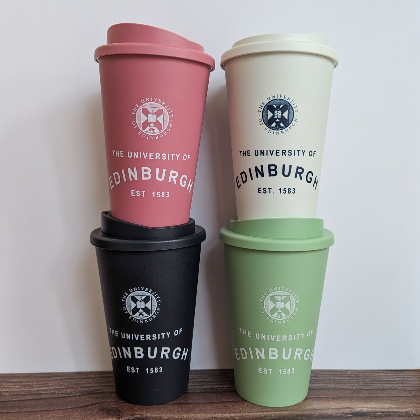 Eco Coffee Cup shown in all four colour options:  Raspberry (Blush Pink), White, Sage Green (Pastel) and Black. Each has the University Crest  and the text 'The University of Edinburgh EST 1583', the word 'Edinburgh' is curved.