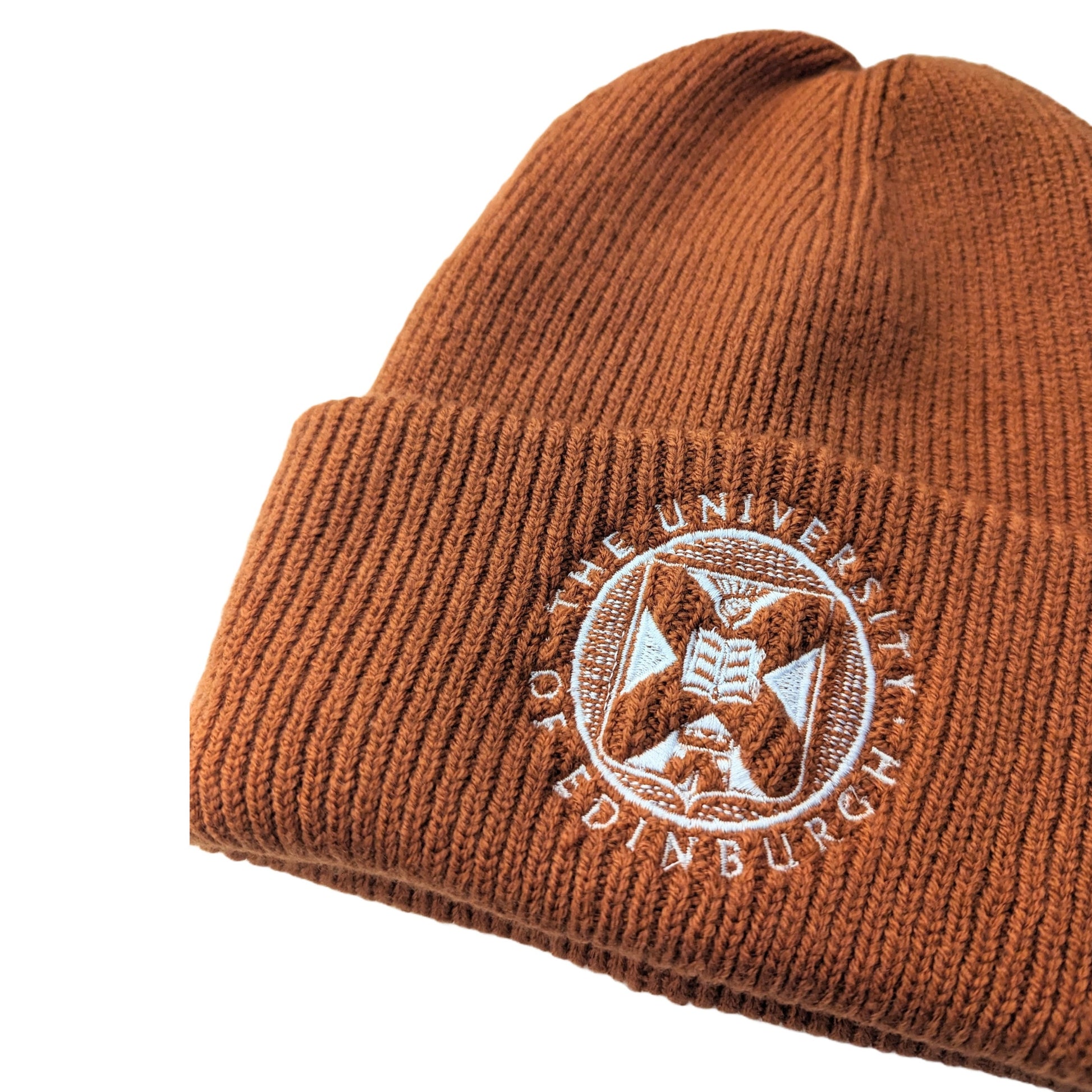 an off-centre close up of rust orange ribbed cotton beanie with white embroidered university crest