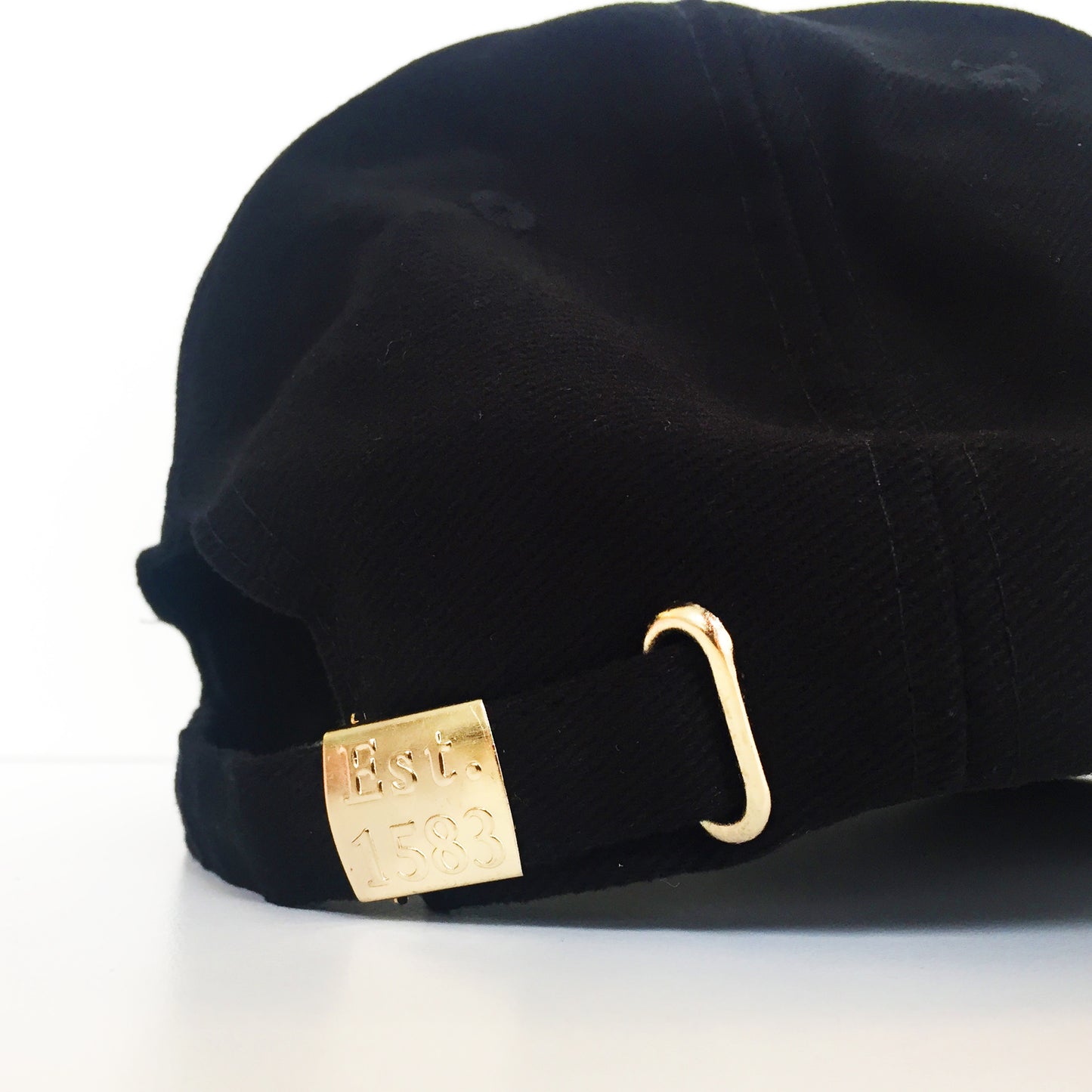 Close up shot of the reverse of a black cap. Adjustable strap with gold hardware that has 'Est. 1583' embossing.