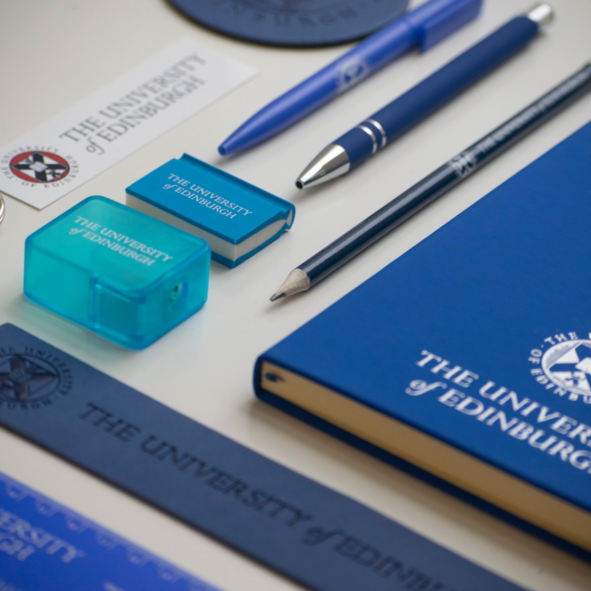 A close up of variety of blue stationery including : A flexible ruler, A leather bookmark, A notebook, A pencil, A soft feel pen ,A recycled, A book Eraser, A pencil sharpener, A leather coaster and a University logo sticker. 