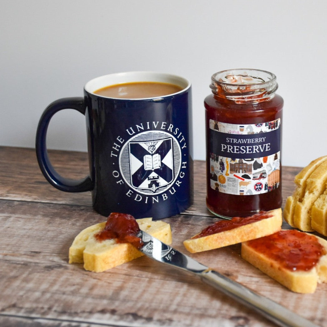 The shiny navy mug with the white University crest on a wooden table is pictured next to our Designed by Esther strawberry preserve. The jar is open, and there is sliced bread with the spread on it.