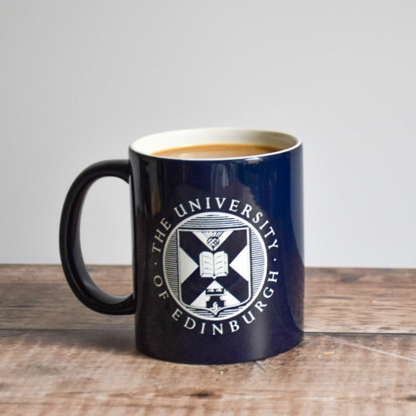 Shiny navy blue mug with a white University crest. There is coffee in the mug, and it is set on a wooden table.