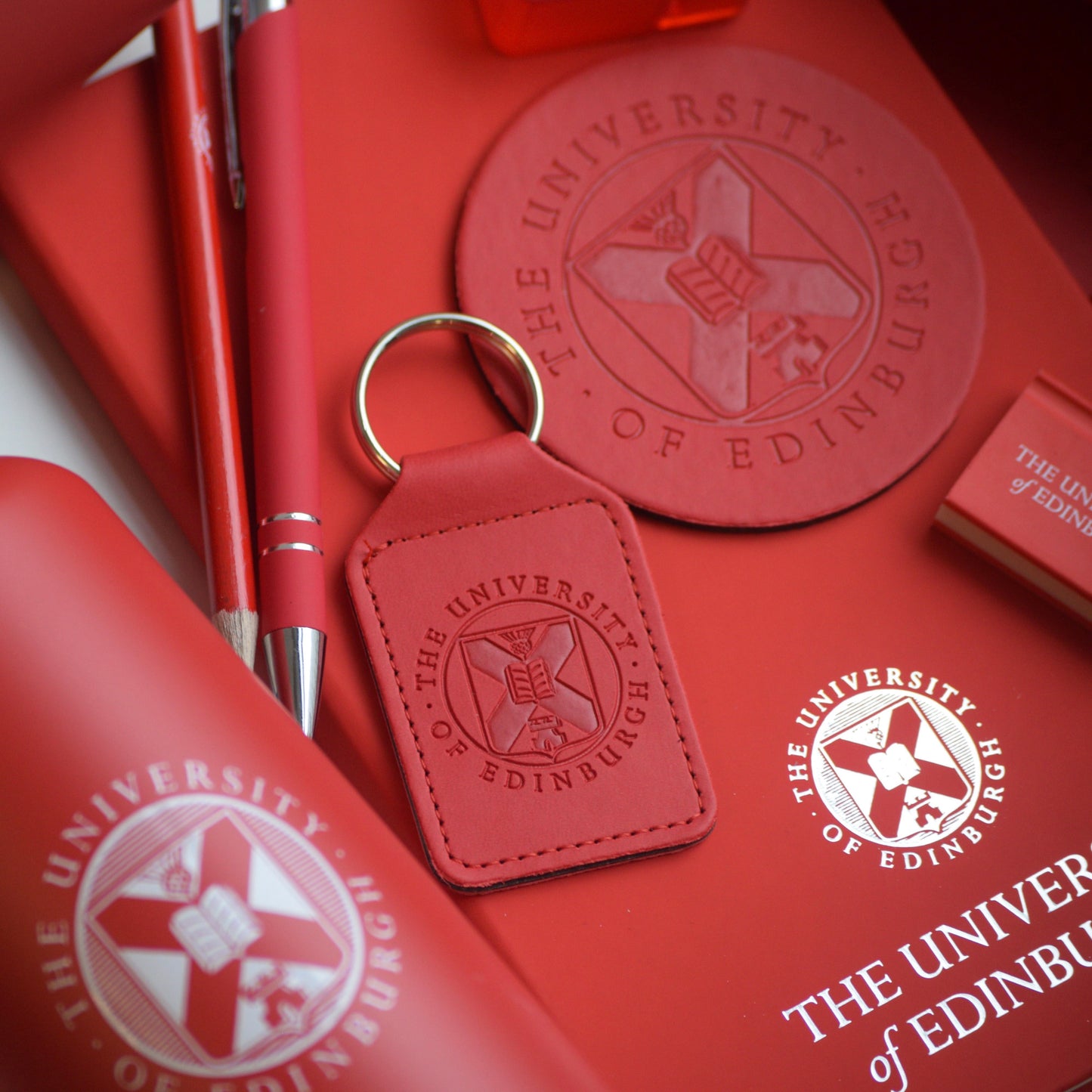 A flat lay of a variety of our red products including : 600ml Water Bottle, Red Pencil, Red Soft Feel Pen, Red Leather Keyring, Red Leather Coaster, Red Book Eraser and Red Foil Crest Notebook. 