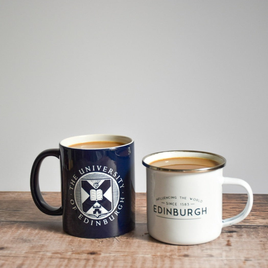 The navy crest mug with the white enamel mug. Both mugs have coffee inside, and are set on a wooden table.