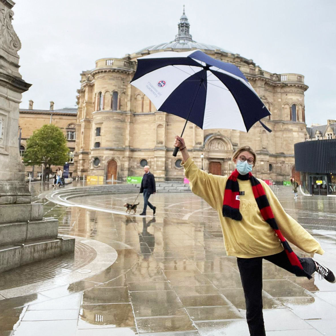 Model in a red and navy striped scarf holing the Large Golf Umbrella outside McEwan hall in the rain. 