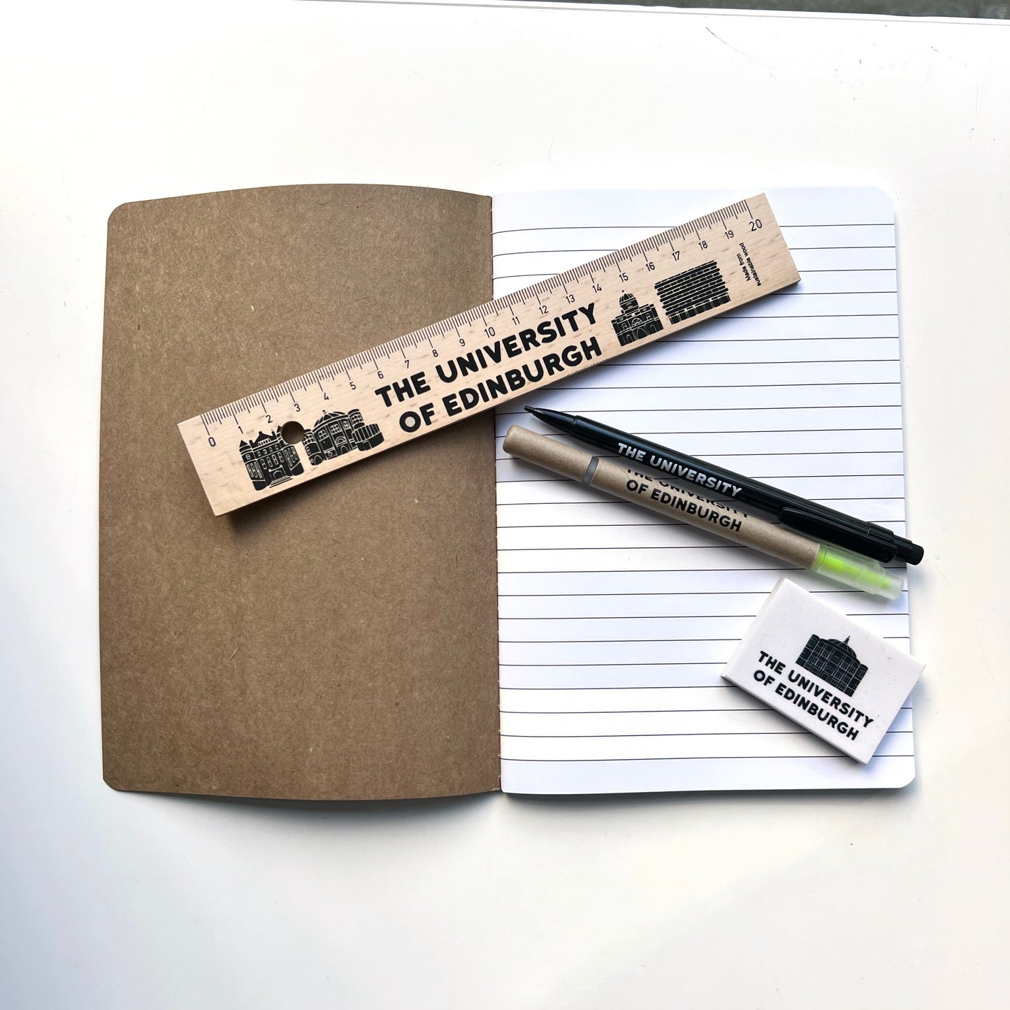 A5 kraft notebook open with lined white pages. Pictured with wooden ruler, mechanical pencil, highllighter twin pen and mcewan hall eraser