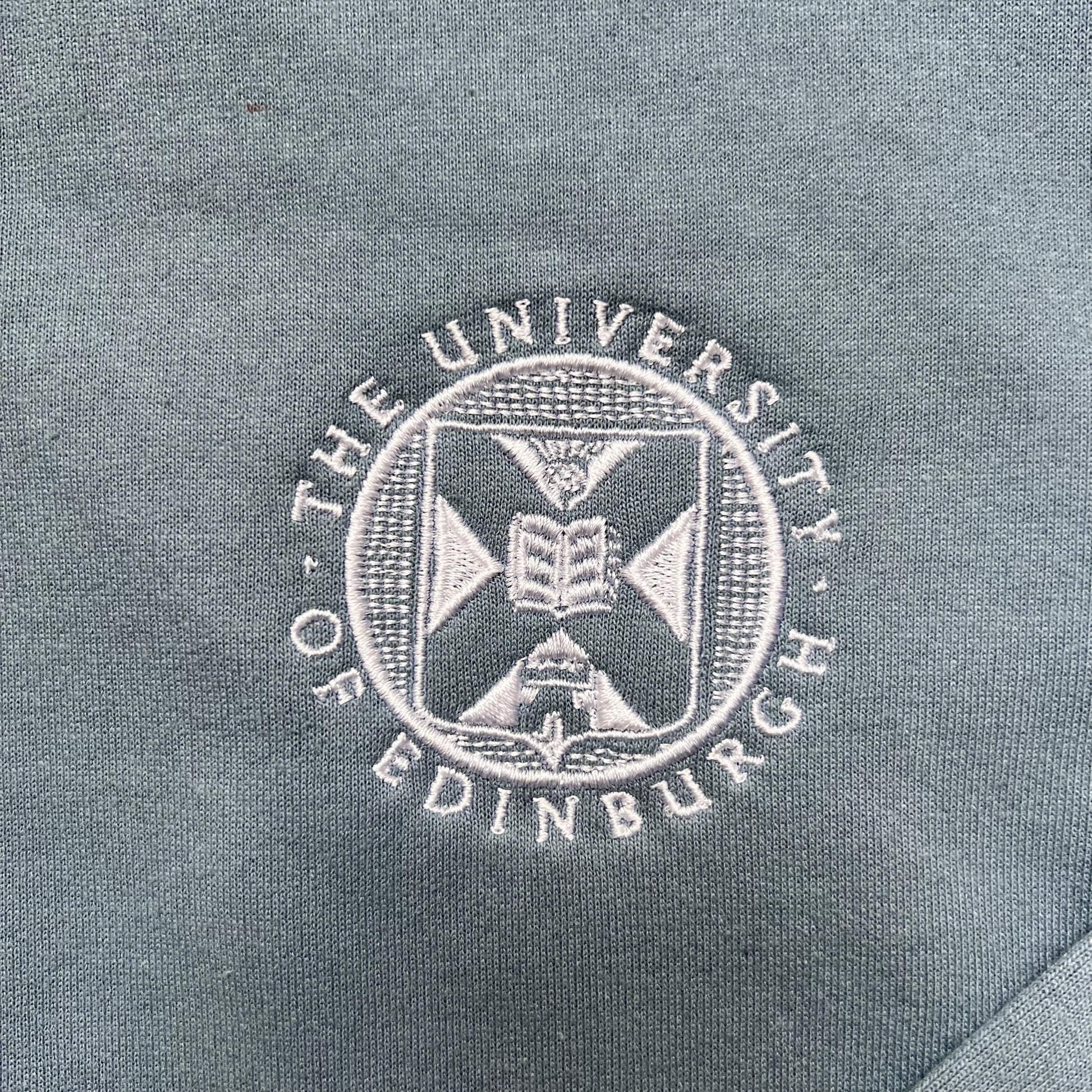 close up of university crest embroidered in white on stone blue sweatshirt 