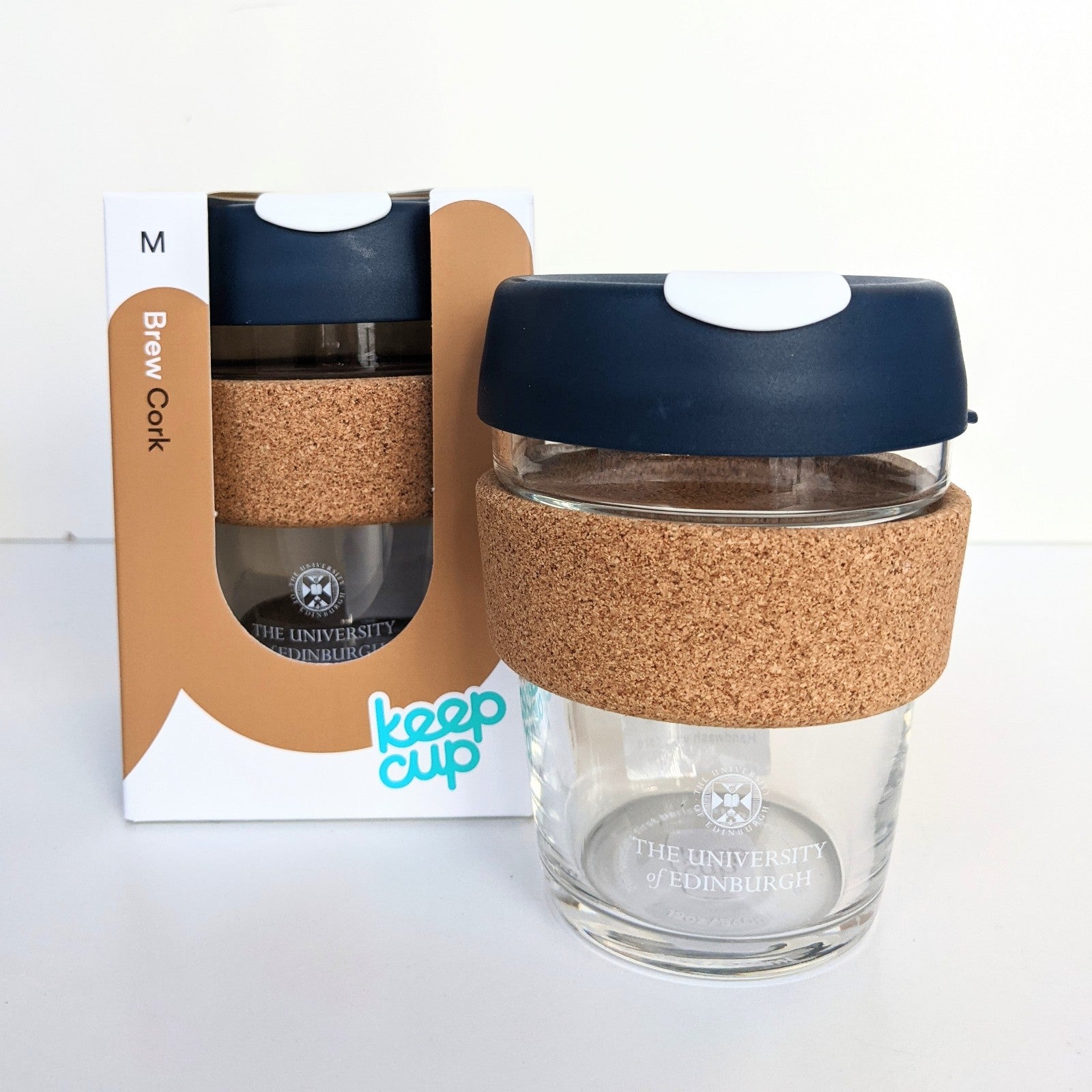 Glass Keep Cup with cork grip and navy blue and white lid displayed beside the same product in its packaging. 