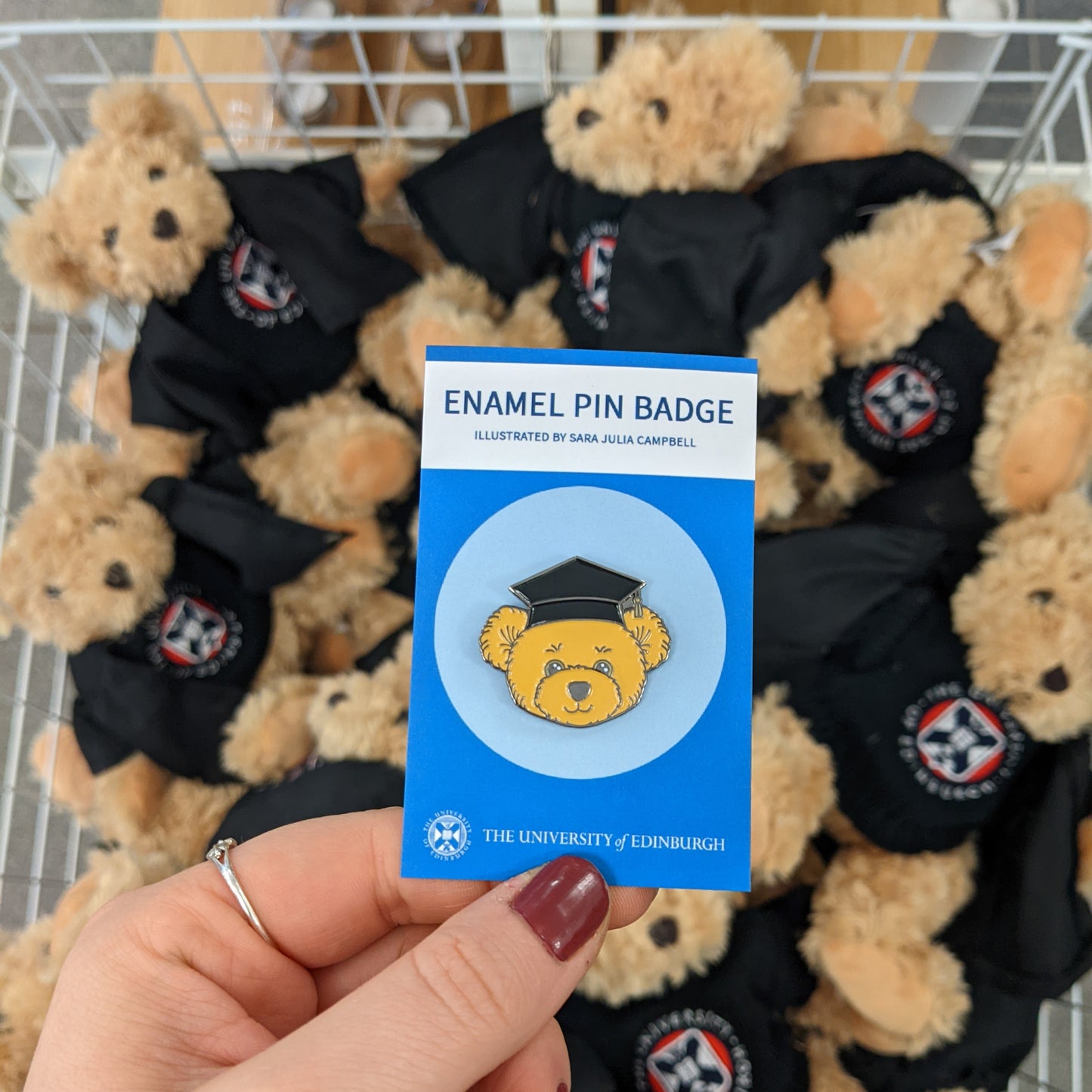 Edinbear in graduation cap pin badge in front of a basket of plush Edinbears 