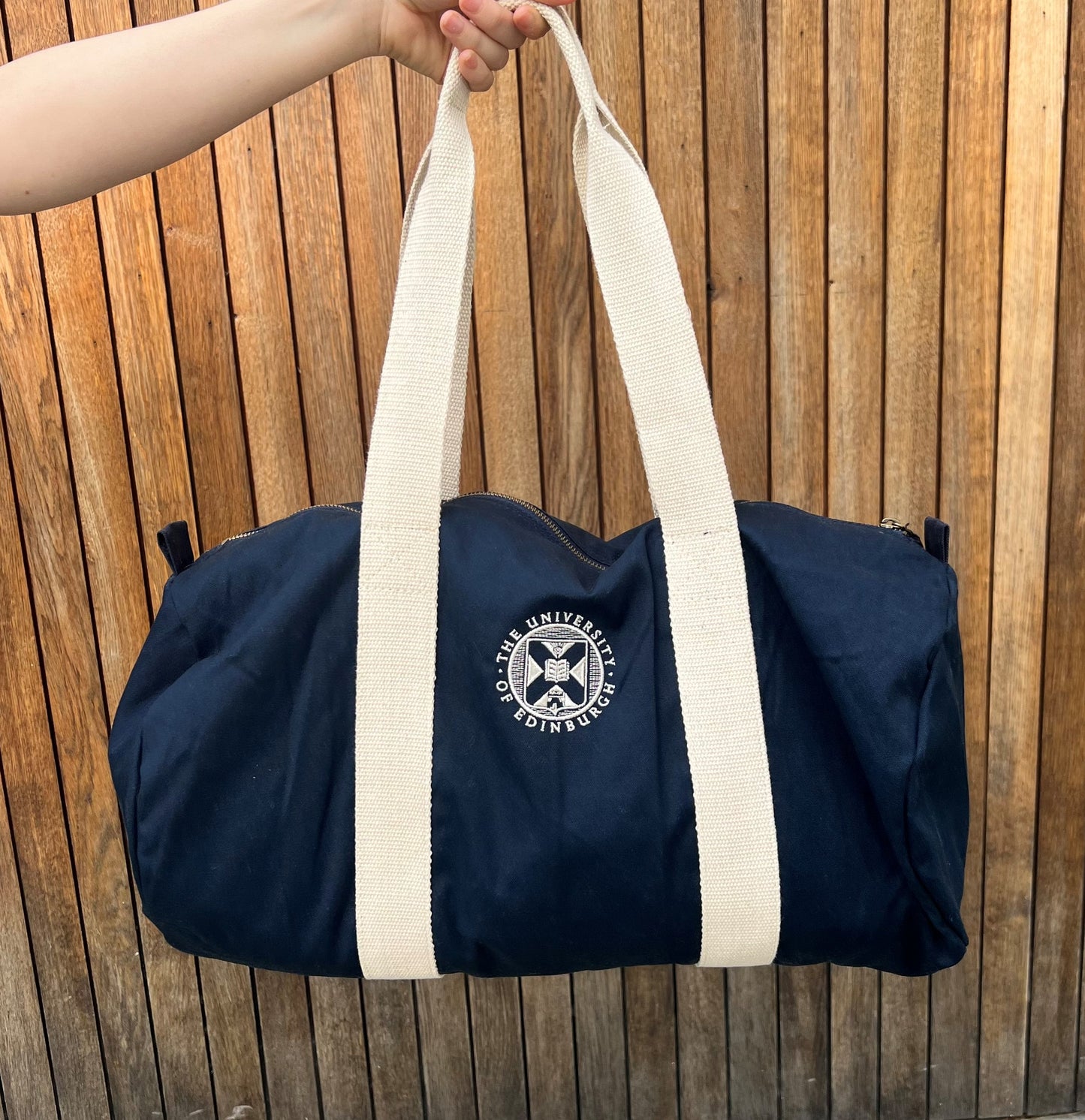 Navy barrel shaped canvas bage with cream handles. University crest is embriodered in the centre. Held up against a wooden fence.