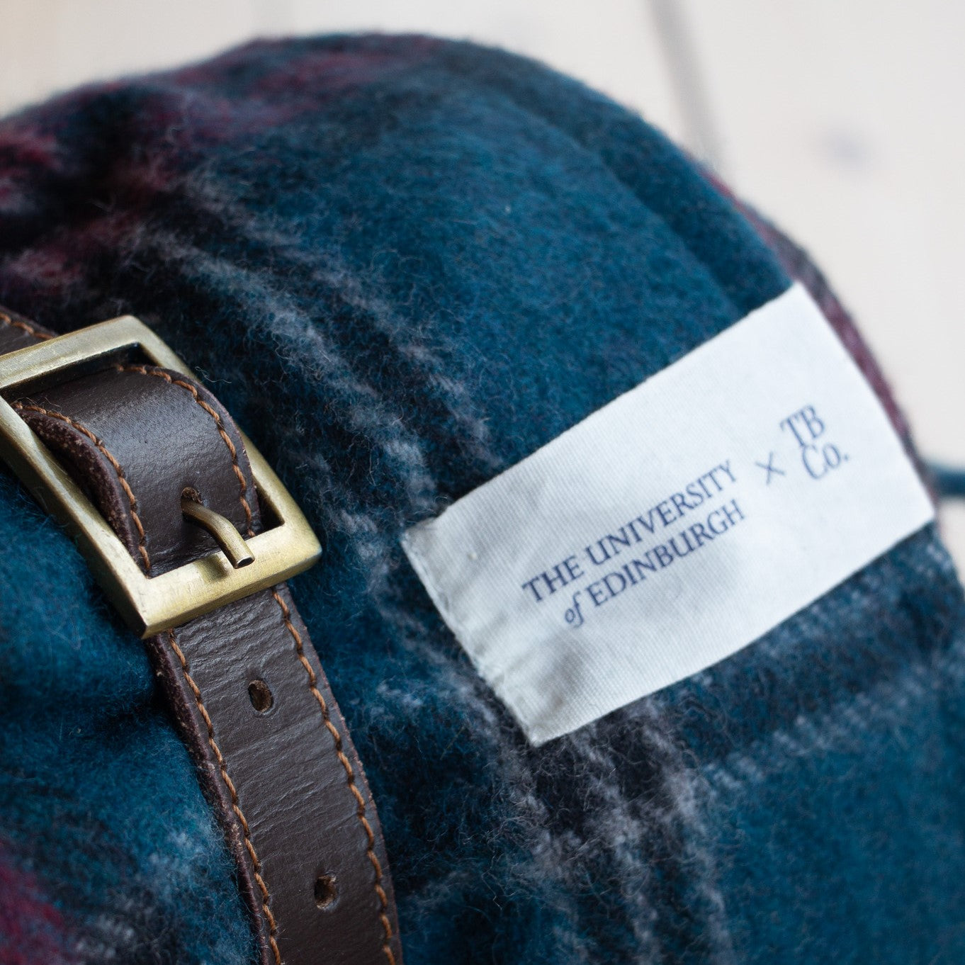 close of up university tartan picnic blanket with leather carrying strap and white 'the university of edinburgh x TBCo.' label