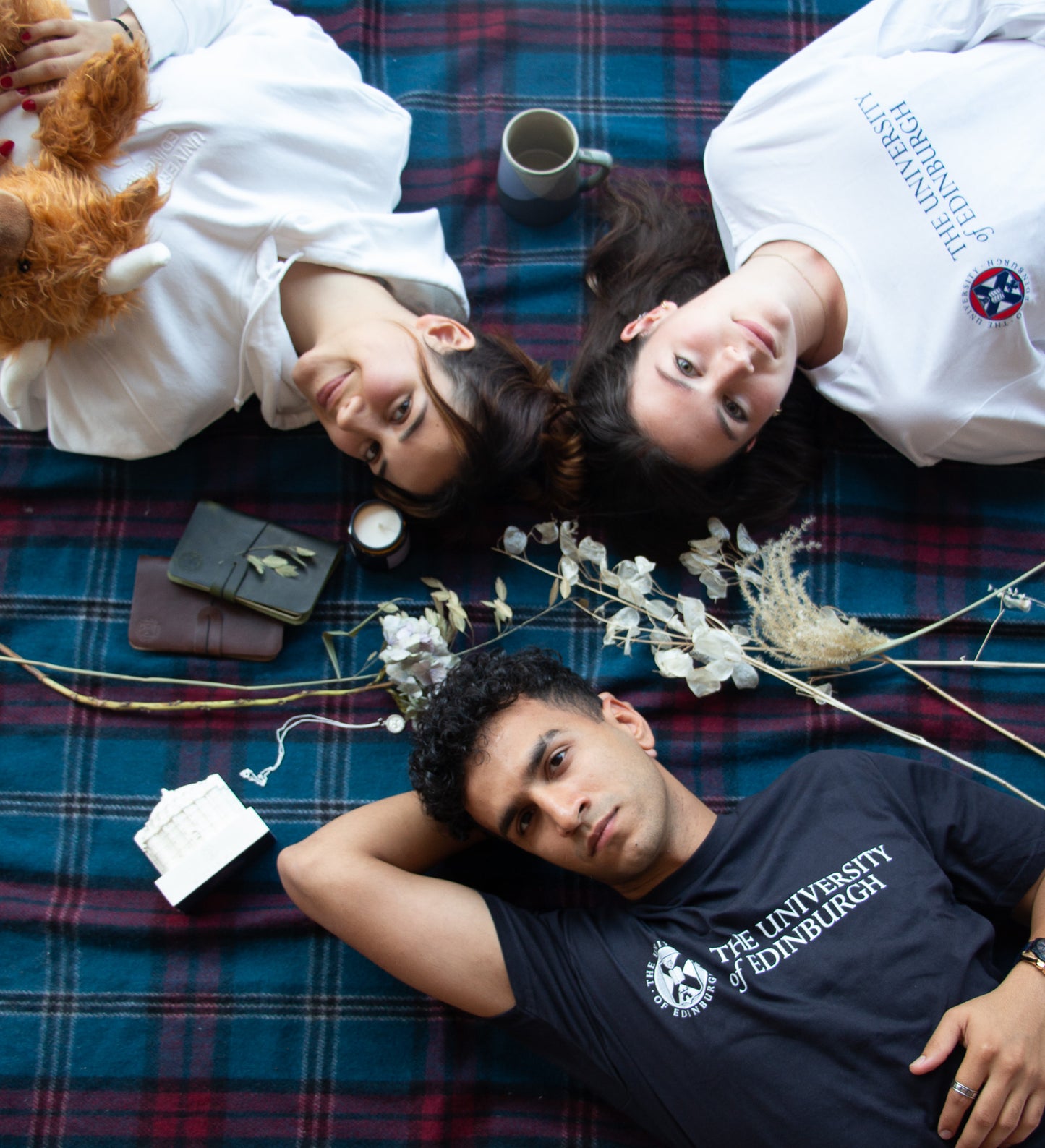 3 models lying on the university tartan picnis blanket with a6 leather note books, flowers and mcewan hall model